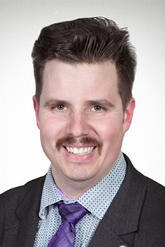 headshot of Dave Elniski a white mail with brown hair and a moustache wearing a dark jacket, checked blue and white shirt and purple tie.
