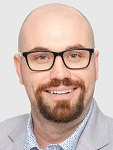 Photo of Adam Donnelly a white male with black-rimmed glasses, beard and moustache wearing a grey jacket and white shirt.